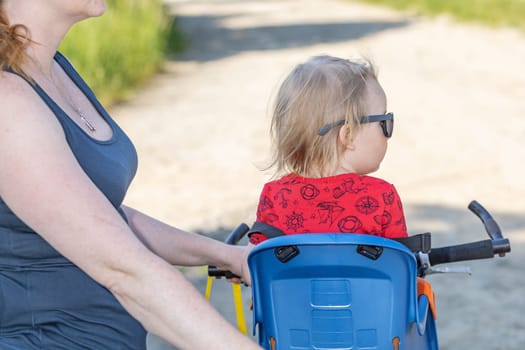 Back view of little boy sitting in bicycle seat without safety helmet. His mother is holding bicycle.