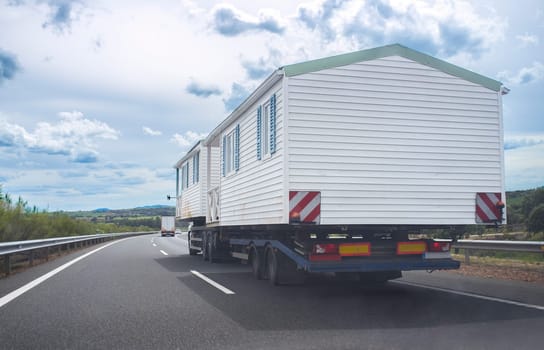 Driving behind heavy-duty truck carrying two prefabricated houses. View from the inside of the car