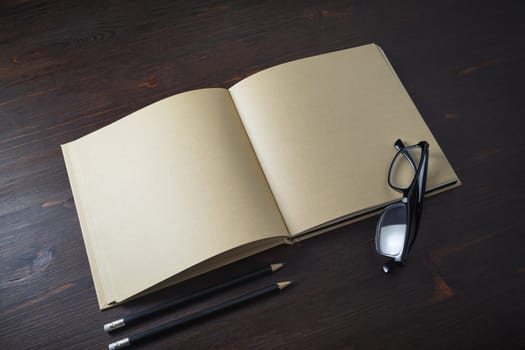 Opened book with blank craft paper pages, pencils and glasses on wooden background.