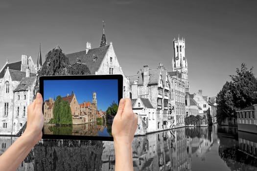 In the bottom left of the photo are hands holding tablet, whose screen contains color photo of the Rozenhoedkaail. Background of the photo contains monochromatic photo of the Old Town of Bruges (Bruges, Belgium)