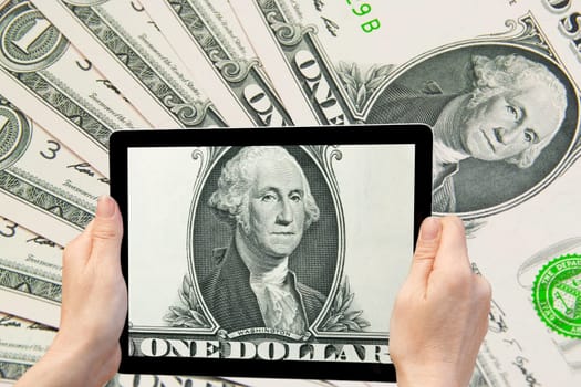 In the bottom right of the photo are hands holding tablet, whose screen contains a closeup view on G. Washington portrait. Background is composed of U.S. one dollar banknotes.