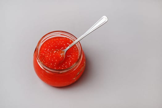 Red caviar in glass jar with spoon on gray background.