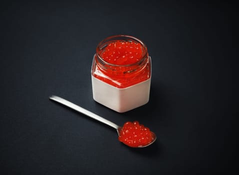 Fresh red caviar, glass jar and spoon on black background.