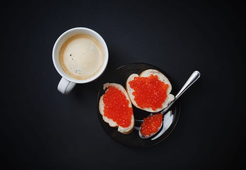 Sandwiches with red caviar, coffee cup, plate and spoon on black background. Top view. Flat lay.