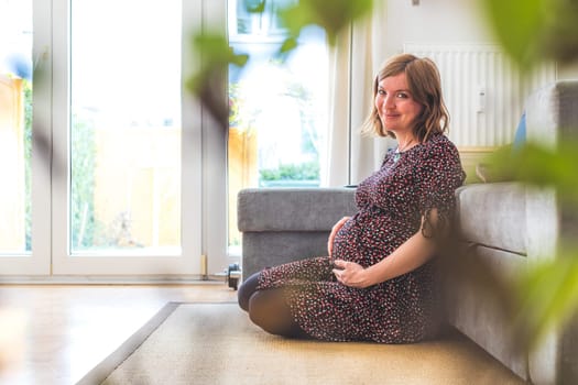 Pregnant Caucasian happy mother is sitting on the floor, touching her tummy, floral dress
