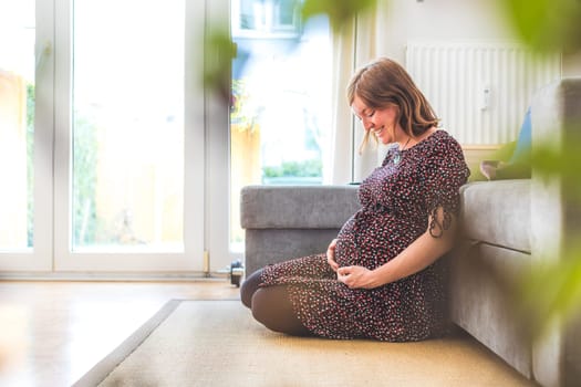 Pregnant Caucasian happy mother is sitting on the floor, touching her tummy, floral dress