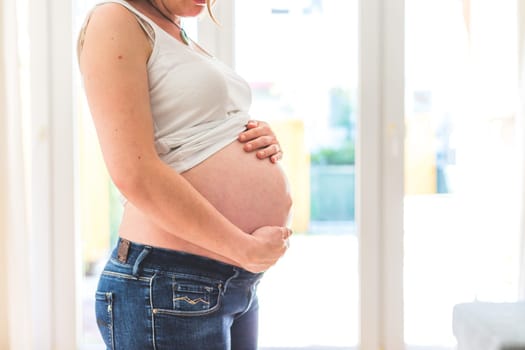 Happy Caucasian pregnant mother touching her naked tummy, blue jeans, close up