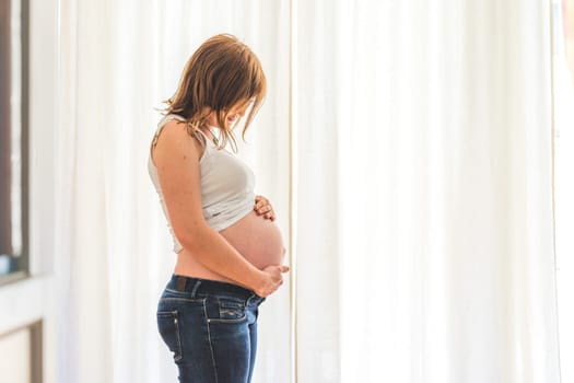 Happy Caucasian pregnant mother touching her naked tummy, blue jeans, close up