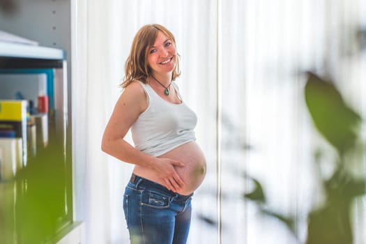 Happy Caucasian pregnant mother touching her naked tummy, blue jeans, close up