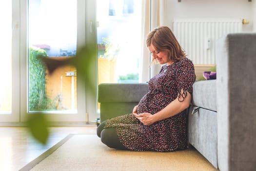 Pregnant Caucasian happy mother is sitting on the floor, touching her tummy, floral dress