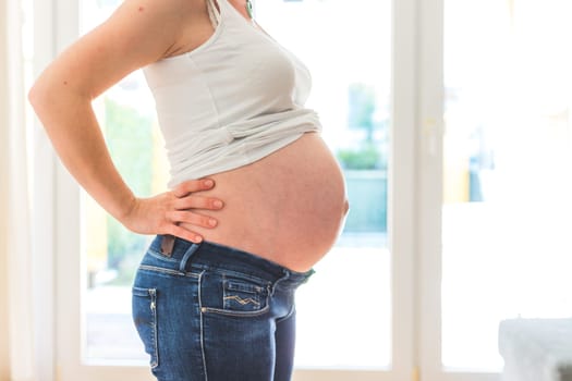 Happy Caucasian pregnant mother touching her naked tummy, blue jeans, close up