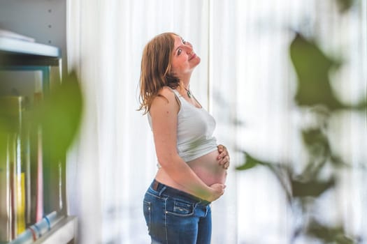 Happy Caucasian pregnant mother touching her naked tummy, blue jeans, close up