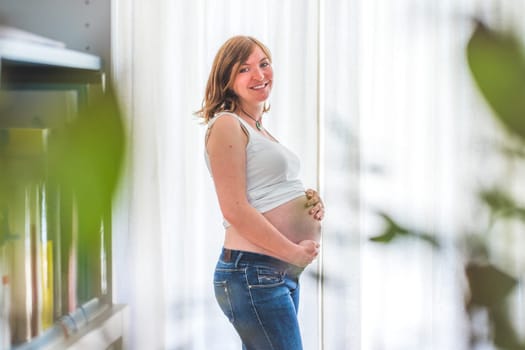 Happy Caucasian pregnant mother touching her naked tummy, blue jeans, close up