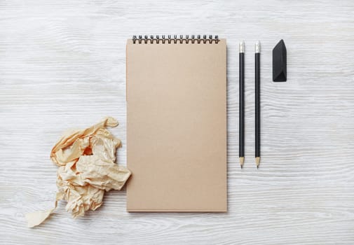 Photo of blank kraft sketchbook, pencil, eraser and crumpled paper on light wooden background. Flat lay.