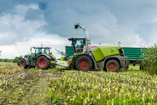 Esens, Germany - October 07, 2020: Side view of two perfectly working together corn harvesters consisting of a corn chopper and a tractor with a tipping trailer under a heavily cloudy sky.
