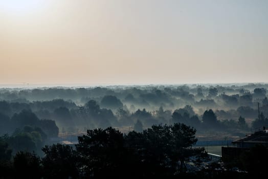 foggy landscape, above deciduous forest, early in the morning. houses in the distance and the surface of the river are visible between the trees. the sun is reflected in the surface of the water. fog to the horizon