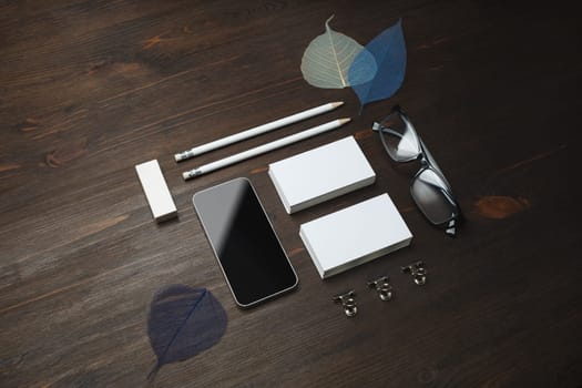 Blank stationery set. Blank business cards, smartphone, pencils, eraser, glasses and leaves on wooden background.