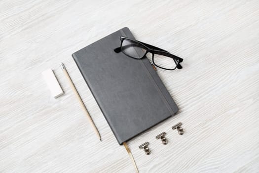 Photo of blank closed notebook, glasses, pencil and eraser on light wood table background. Responsive design mockup. Stationery elements.