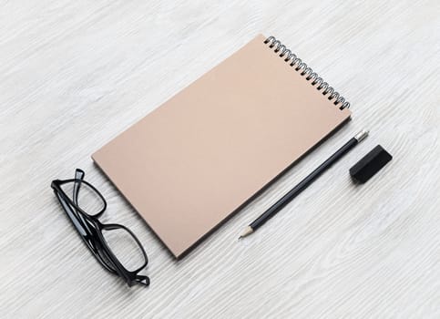 Blank stationery set. Kraft sketchbook, glasses, pencil and eraser on light wooden background. Template for placing your design. Responsive design mockup.