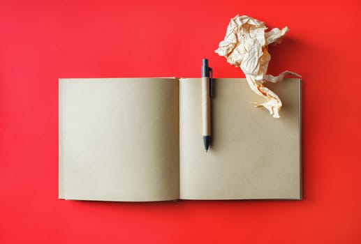 Photo of kraft open book with blank pages, crumpled paper and pen on red paper background. Mockup for placing your design. Top view. Flat lay.