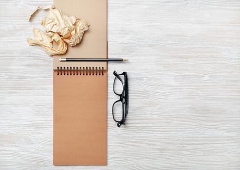 Opened notebook with blank craft paper pages, glasses, pencil and crumpled paper on light wood table background. Flat lay.
