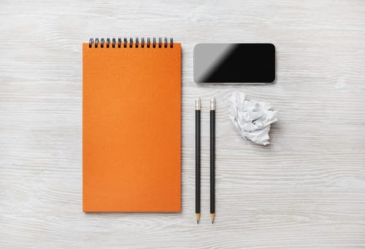 Blank orange notepad, smartphone, pencils and crumpled paper on light wood table background. Top view. Flat lay.