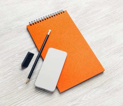 Blank orange notebook, smartphone, pencil and eraser on light wooden background. Stationery mock up.