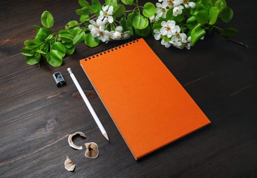 Photo of blank stationery template. Orange sketchbook, pencil, sharpener and spring flowers on wood table background.
