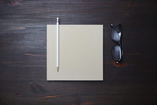 Blank closed book, pencil and glasses on wood table background. Stationery template. Flat lay.