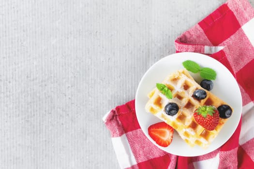 White plate on red kitchen towel with stack homemade Belgian waffles, blueberry, strawberry cuts, mint leaf, sugar powder on gray background