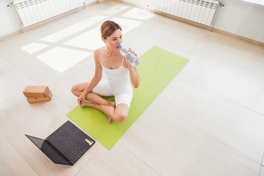 Woman sitting on mat in sportswear, drinks water and doing sport at home with laptop online
