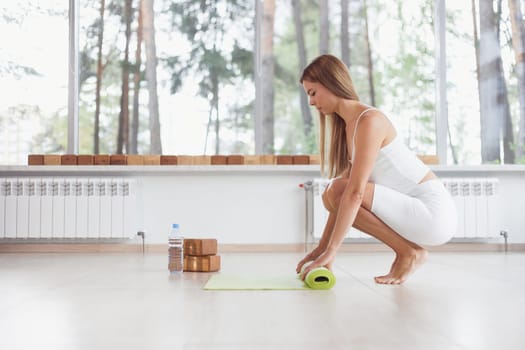 A barefoot woman twists a purple yoga mat and fitness on the parquet floor.