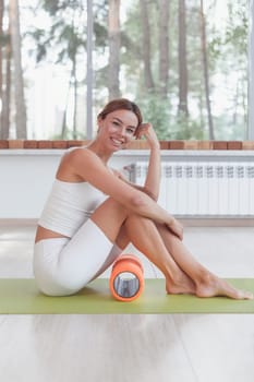 Sporty woman with foam roller sitting on mat in sport hall