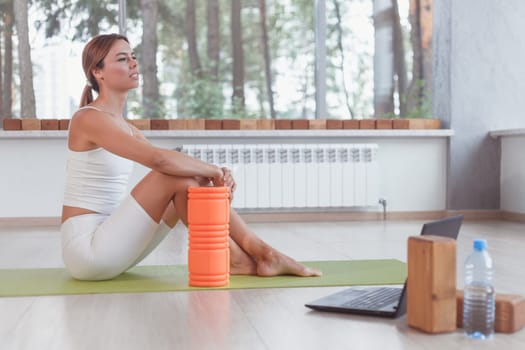 Sporty woman with foam roller sitting on mat in sport hall