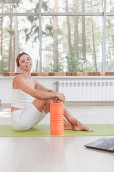 Sporty woman with foam roller sitting on mat in sport hall