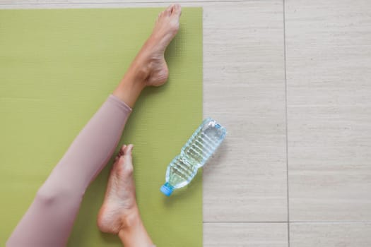 Woman in sportswear sitting on gymnastic mat with bottle water. Flat lay, close up view on legs, topo view.
