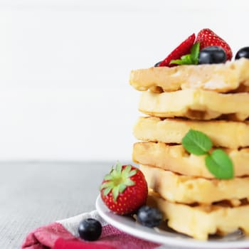 White plate on red kitchen towel with stack homemade Belgian waffles, blueberry, strawberry cuts, mint leaf, sugar powder on gray background