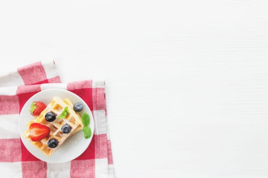 White plate on red kitchen towel with stack homemade Belgian waffles, blueberry, strawberry cuts, mint leaf, sugar powder on gray background