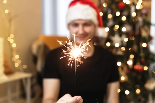 Young man in Santa hat is holding burning sparkler near Christmas tree. Christmas lights in guy hands on blurred background with decorated Christmas tree in living room. Smiling man looking at fire