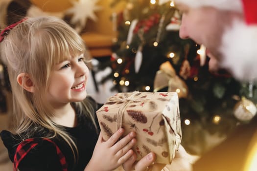 Young father in red Santa hat giving Christmas gift to his smiling daughter near decorated Christmas tree. Girl dressed in festive red-black Christmas outfit. They smiling, happy because of New Year