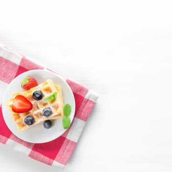 White plate on red kitchen towel with stack homemade Belgian waffles, blueberry, strawberry cuts, mint leaf, sugar powder on gray background