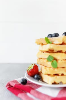 White plate on red kitchen towel with stack homemade Belgian waffles, blueberry, strawberry cuts, mint leaf, sugar powder on gray background
