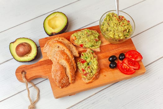 Freshly prepared guacamole in small glass bowl, bread, tomatoes, olives at working board and two avocados halves next, view from above.