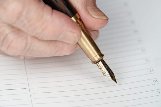 Gold coloured fountain ink pen in senior woman hand about to write something on empty daily planner page, closeup detail.