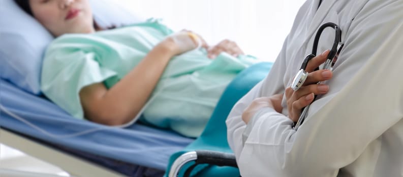 Male doctor with crossed arms holding stethoscope and blurred background of woman patient lying on bed in hospital room. Healthcare and insurance concept.