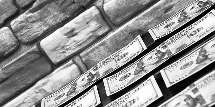 Dollars in one-hundred-dollar bills are spread out on a table near the brick wall. The dollars are arranged in a row. US dollars. On the brown brick wall there is a place for an inscription, a logo. Black and white style. Layout, template, mockup.