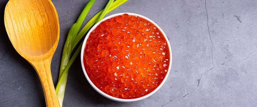 Red caviar in a wooden cup on a gray background with a spoon and green onions. A large pile of bright caviar. Fresh delicious caviar. A place for advertising, logo, label, layout, mockup. Layout, template.