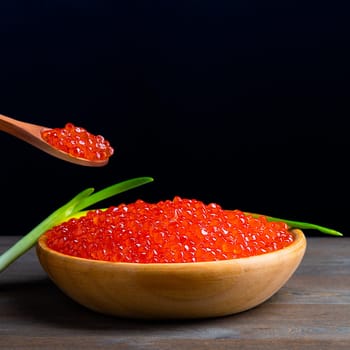 Red caviar in a wooden cup on a black background with a spoon and green onions. A large pile of bright caviar. Fresh delicious caviar. A place for advertising, logo, label, layout, mockup. Layout, template.