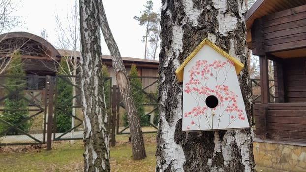 Ukraine, Kiev - April 17, 2020. Beautiful white painted birdhouse made of wood on a birch in the forest near a brown country wooden house. Homemade bird feeder. Art birdhouse on a tree in the garden