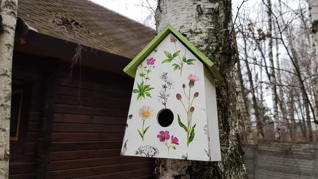 Ukraine, Kiev - April 17, 2020. Beautiful white painted birdhouse made of wood on a birch in the forest near a brown country wooden house. Homemade bird feeder. Art birdhouse on a tree in the garden